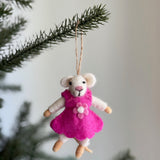 A handmade felt mouse ornament, an individual piece from the Mouse Family Ornament set, hangs by a jute string on a pine branch against a soft white background. This charming felted mouse features a white head, pink nose, and small black eyes. It wears a bright pink dress adorned with a delicate flower, creating a whimsical and festive atmosphere.