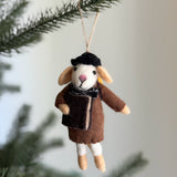 A handmade felt mouse ornament hangs from a pine branch, its jute string suspending it against a soft, neutral backdrop. The charming felt mouse wears a brown coat, black hat, and a black and white patterned scarf. Its white felt face features small black eyes and a pink nose, showcasing its whimsical design.