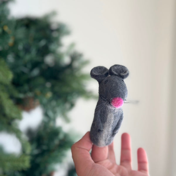 A handcrafted felt mouse finger puppet is presented by a female hand against a festive, blurred Christmas tree background. This charming gray mouse puppet features rounded ears, a prominent pink nose, and delicate whiskers, showcasing its soft texture. Designed for imaginative play and storytelling, this unique felt toy adds a whimsical touch to festive celebrations.