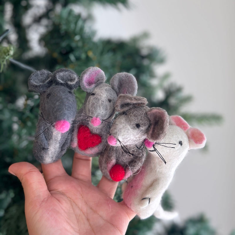 A handmade felt mouse finger puppet is gently presented by a female hand, set against a soft, blurred Christmas tree backdrop. This charming felt toy for kids features pointed ears with pink inner details, black thread whiskers, and small black eyes, evoking a sense of playful creativity.