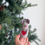 A handmade needle felted mouse finger puppet, holding a red heart, is gently held by a female hand against a blurred Christmas tree background. This gray 100% wool mouse features pink inner ears, a pink nose, and whiskers. This charming, fair-trade felt puppet from Nepal is crafted for imaginative play.