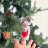 A charming handmade needle felted mouse finger puppet, holding a bright red heart, is presented by a female hand. The gray wool felted mouse features pink inner ears, a tiny pink nose, and delicate black whiskers, showcasing its unique, handcrafted design. This delightful kid's finger puppet is set against a softly blurred Christmas tree background.