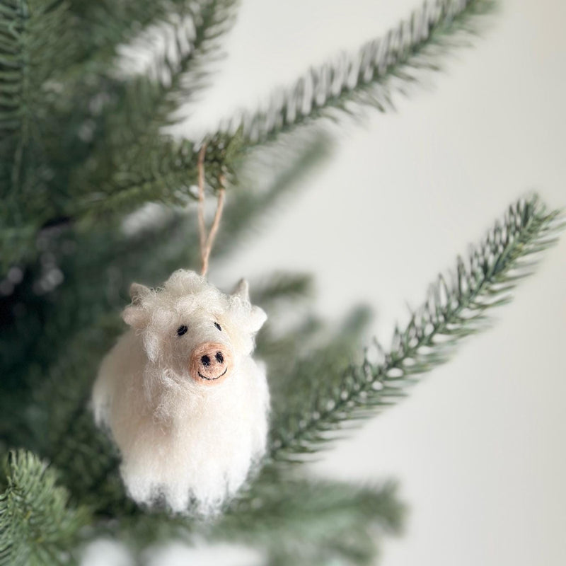 A fluffy white needle felted Highland Cow Christmas ornament hangs from a green Christmas tree branch. The soft ornament features small horns, a smiling face, and is suspended by a jute string, creating a joyful, festive mood.