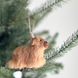 A charming Needle Felted Highland Cow Ornament is prominently displayed on a vibrant green Christmas tree branch. This handmade felt ornament features a fluffy, light brown body with gray horns and delicate needle-felted details, complete with a jute string for hanging.