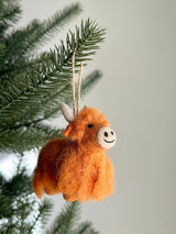A needle felted Highland cow ornament hangs from a green pine needle branch on a Christmas tree. This unique Christmas ornament features fluffy orange wool, small black eyes, light gray horns, and a white circular muzzle with a stitched black smile, suspended by a jute string.