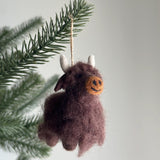 A needle felted Highland Cow ornament, crafted from soft brown wool felt, hangs from a green pine branch against a calm white background. This charming Highland Cow features cute white horns, small ears, and delicate needle-felted details, making it a delightful Christmas tree ornament.