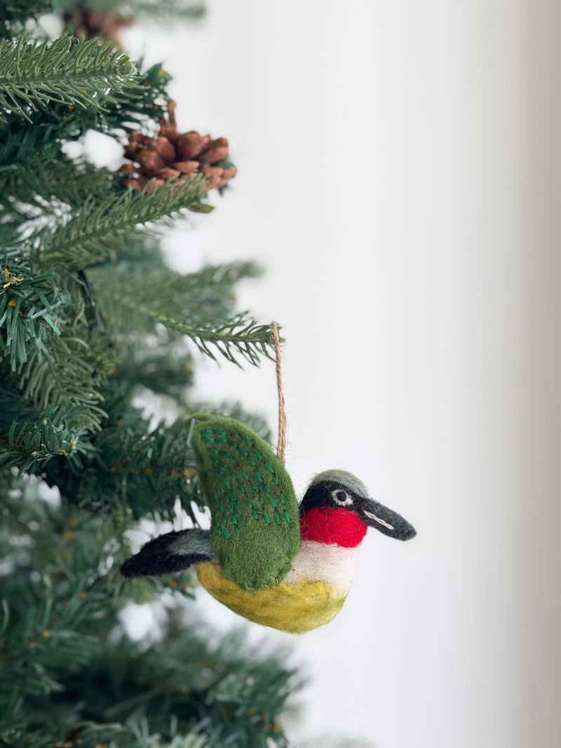 A needle felted hummingbird ornament hangs delicately from a jute string on a lush green Christmas tree, adorned with pine needles and a few brown pine cones. The handcrafted felt hummingbird features a body transitioning from yellow to white, a vibrant red throat, and a black beak. Its green wings are accented with decorative green stitching, adding texture.