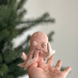 A light pink wool felt octopus finger puppet is showcased on a human hand, set against a softly blurred Christmas tree background. This handmade octopus features eight tiny curled tentacles, small poppy eyes, and a charming stitched grin. Its cheerful, handcrafted design emphasizes its unique charm, perfect for imaginative play.