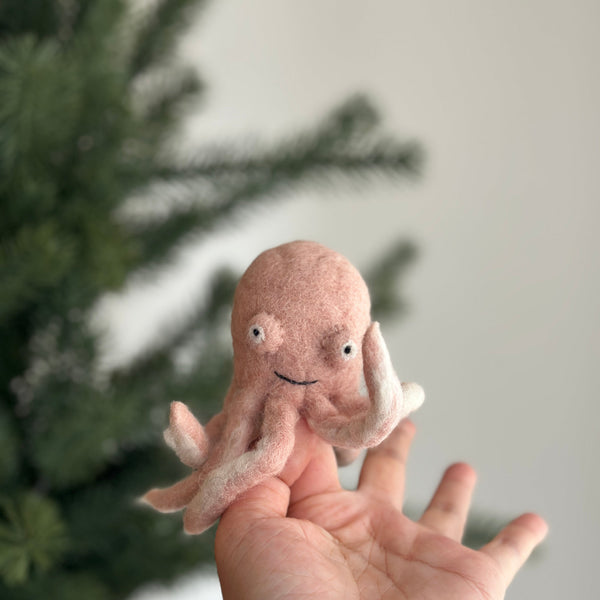 A light pink wool felt octopus finger puppet is showcased on a human hand, set against a softly blurred Christmas tree background. This handmade octopus features eight tiny curled tentacles, small poppy eyes, and a charming stitched grin. Its cheerful, handcrafted design emphasizes its unique charm, perfect for imaginative play.