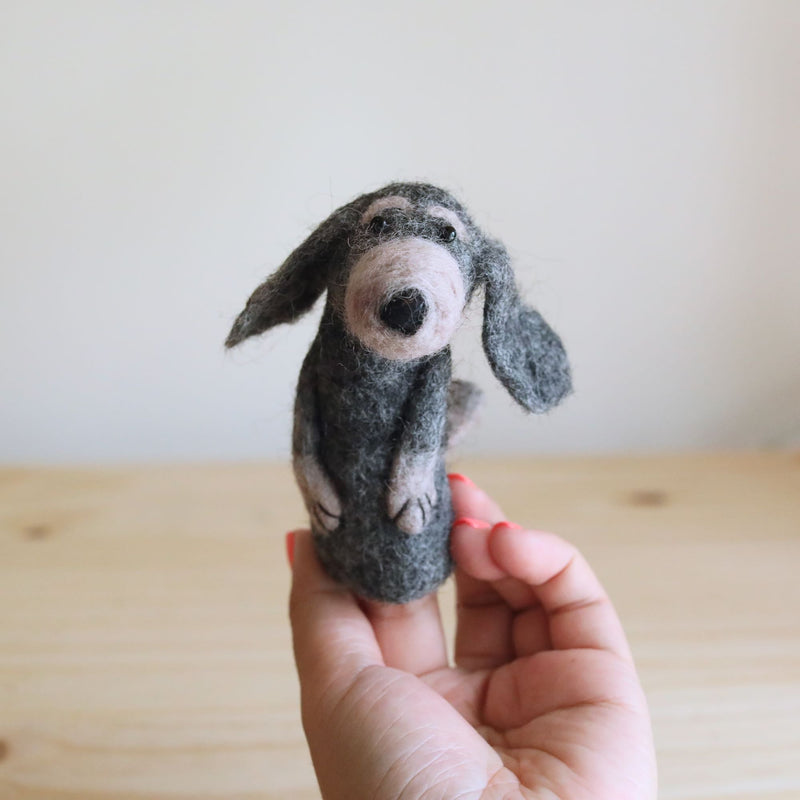 A woman's hand with red nail polish gently holds a charming needle felted old gray dog finger puppet against a light wooden surface. This 3D handmade wool felt dog puppet features gray fur, a light pink nose, and long, droopy ears, reflecting its artisanal fair-trade craftsmanship.