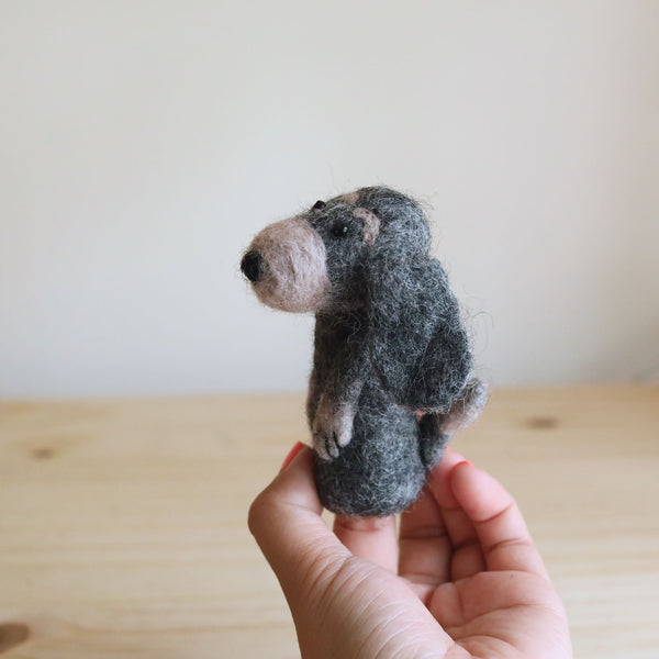 A handmade needle felted old gray dog finger puppet is held by a female hand, displayed against a soft white wall and a wooden surface. This unique wool felt finger puppet features a gray body, light pink nose, and visible black eyes and arms, reflecting its charming craftsmanship. It's an ideal felt puppet for finger play, showcasing detailed design.