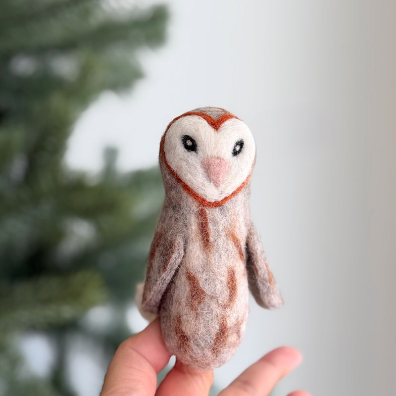 A handmade wool felt owl finger puppet is gently held in a female hand, presented against a softly blurred Christmas tree background. This felted finger puppet features a distinctive white heart-shaped face framed by reddish-brown markings, and a small pink beak. Its textured wool felt body is adorned with brown stripes, giving the owl a realistic, feathered appearance. The simple design and soft colors create a whimsical, comforting mood.