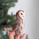 A wool felt owl finger puppet is held by a hand, presented against a soft-focus Christmas tree background. This handcrafted owl puppet features a distinctive heart-shaped face with a reddish-brown outline, white center, small pink beak, and black eyes. Its wool felt body displays a gradient of browns and creams, resembling natural owl plumage, complete with small felted wings and tail.
