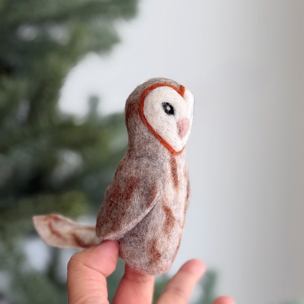 A wool felt owl finger puppet is held by a hand, presented against a soft-focus Christmas tree background. This handcrafted owl puppet features a distinctive heart-shaped face with a reddish-brown outline, white center, small pink beak, and black eyes. Its wool felt body displays a gradient of browns and creams, resembling natural owl plumage, complete with small felted wings and tail.