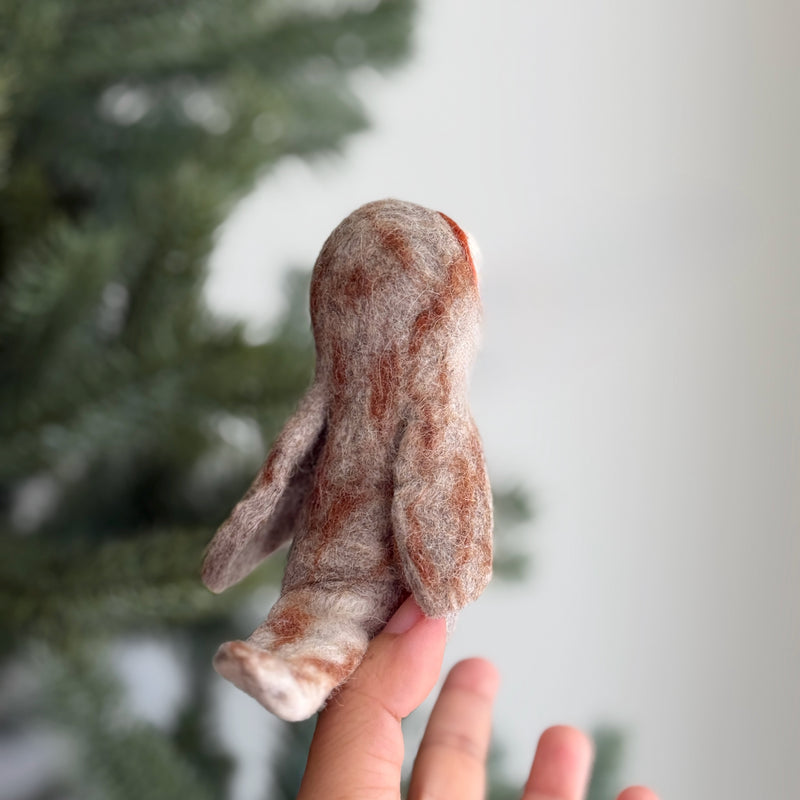 A handcrafted wool felt owl finger puppet is displayed against a blurred background. This unique kids felt toy is made with needle-felted wool, featuring a mottled pattern of light browns and tans, and small wings extending from its sides. Its soft texture highlights the intricate 3D design.