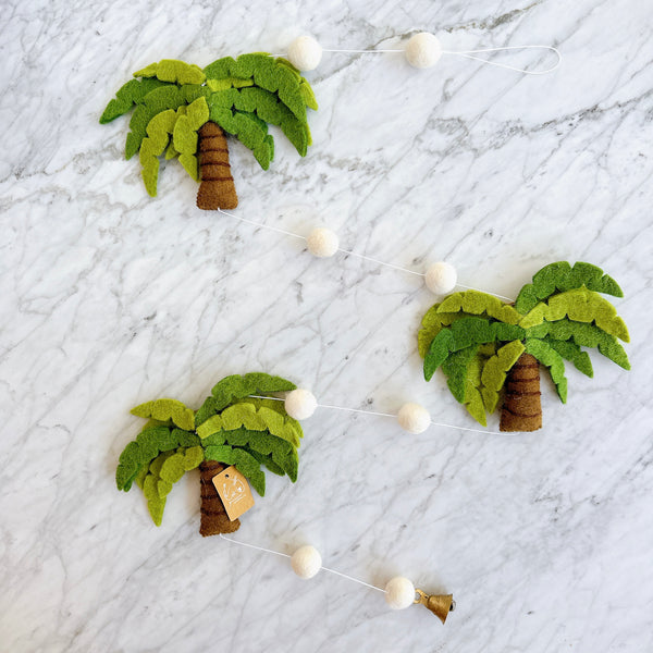 A felt palm tree garland rests on a marble backdrop, creating a tropical and whimsical scene. This decorative garland features three felt palm trees, each with a brown trunk and layered green fronds. They are connected by evenly spaced white felt balls. A small copper bell hangs delicately from one end of the garland.