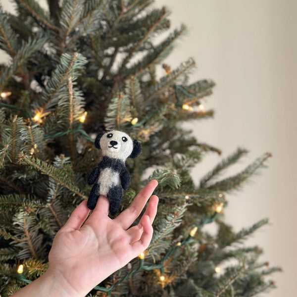 A charming handmade panda finger puppet is playfully presented by a hand, set against a blurred background featuring a festive Christmas tree adorned with small lights. This endearing wool felt panda, a unique felt toy for kids, showcases its classic black and white coloring, a cute round face, plump belly, black ears, and a black nose.