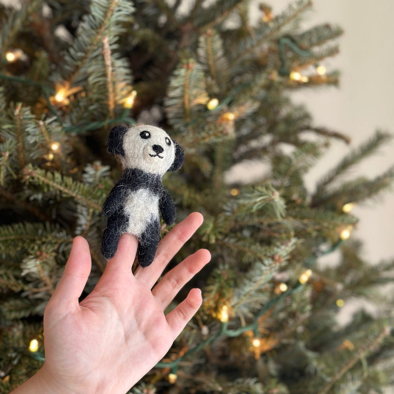 A handcrafted felt panda finger puppet is held upright against a warm, lit Christmas tree. This black and white panda features a round face, plump body, black ears, and eye markings that distinctively contrast with its white face and belly. The charming felt toy is ready for imaginative play.