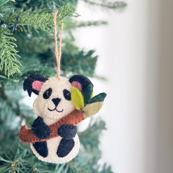 A charming wool felt panda ornament hangs on a Christmas tree. The cream-colored panda features black markings around its eyes, on its ears, and on its lower body, and is depicted holding a green bamboo stalk. This handcrafted felt ornament, made from cozy wool felt, includes a jute thread loop for easy hanging, evoking a playful holiday spirit.
