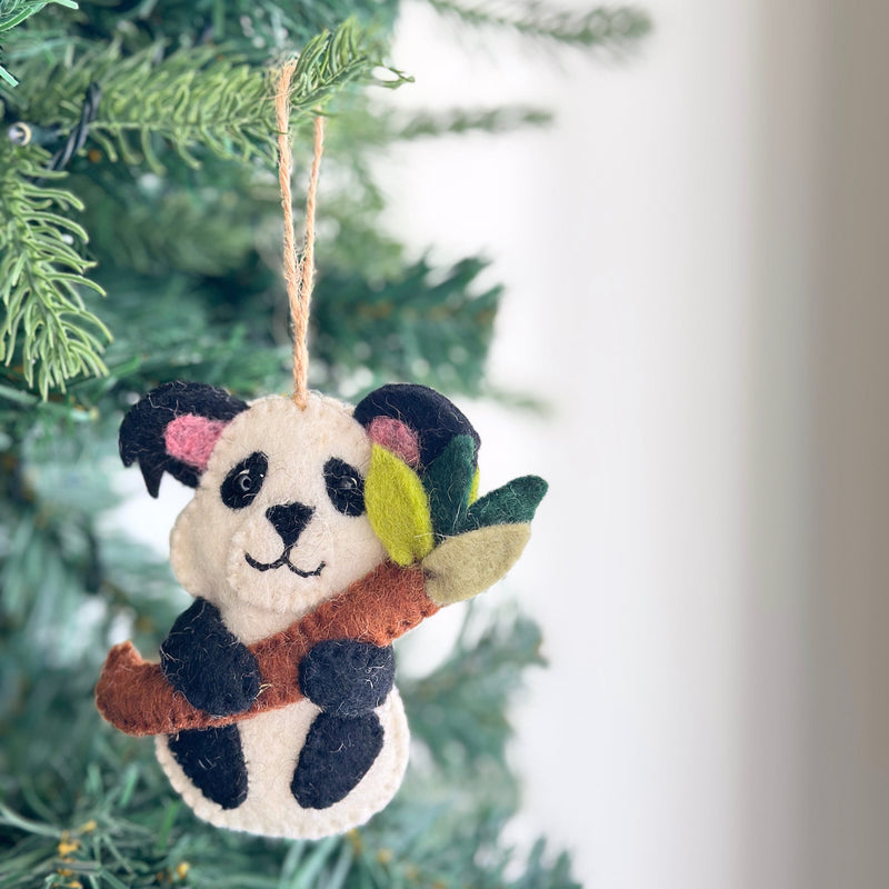 A charming wool felt panda ornament hangs on a Christmas tree. The cream-colored panda features black markings around its eyes, on its ears, and on its lower body, and is depicted holding a green bamboo stalk. This handcrafted felt ornament, made from cozy wool felt, includes a jute thread loop for easy hanging, evoking a playful holiday spirit.