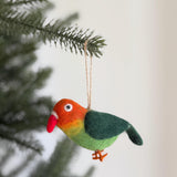 A vibrant wool felt parrot ornament hangs from a Christmas tree branch against a soft white backdrop. This unique Christmas ornament features a colorful gradient of orange, yellow, and green on its body, dark green wings, a bright red beak, and orange feet. The handcrafted felt ornament boasts textured wool and a jute string for hanging, creating a festive and cheerful mood.