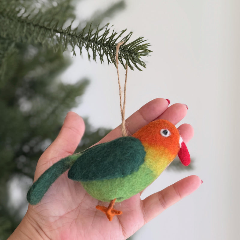 A vibrant wool felt parrot ornament hangs on a blurred Christmas tree, creating a festive atmosphere. This unique Christmas ornament displays a color gradient from orange on its head to yellow and green on its body, complemented by a red beak and orange feet. Crafted from textured wool felt, it includes a jute string for easy hanging.