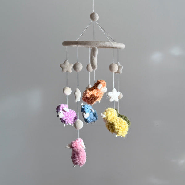 A soothing pastel sheep baby mobile, handcrafted from soft wool felt, hangs gently in front of a white background. The mobile features several pastel-colored sheep suspended from a felt hoop, arranged in a circular pattern around the hoop. Each sheep varies in size and adds to the calming atmosphere of the scene.