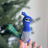 A vibrant handcrafted needle-felted peacock finger puppet is held by a female hand, set against a soft background with a blurred Christmas pine tree. Made from soft wool felt, this ethically crafted peacock puppet displays vibrant blue and green hues with gray accents and a mini fan tail featuring delicate hand-stitched details.