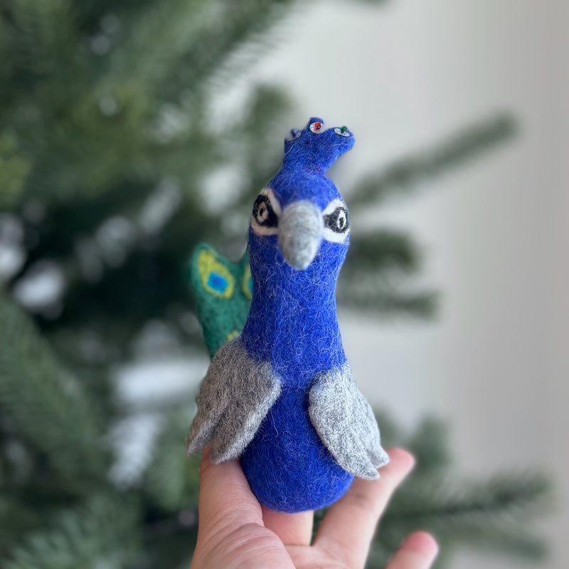 A handcrafted needle-felted peacock finger puppet is held gently in a female hand, prominently displayed against a blurred Christmas tree and soft white wall background. This unique wool felt finger puppet features a predominantly blue body with a grey beak, grey wings, and a distinctive green and blue tail fan, showcasing its ethical craftsmanship.