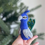 An ethically made, needle-felted peacock finger puppet is gently presented by a female hand against a blurred Christmas tree background. This handcrafted wool felt peacock features a vibrant blue body, gray wings, and a beak. Its distinctive green and blue mini fan tail highlights the exquisite craftsmanship and playful nature of this charming felt finger puppet.