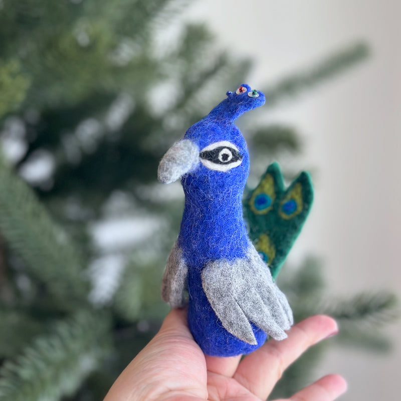 An ethically made, needle-felted peacock finger puppet is gently presented by a female hand against a blurred Christmas tree background. This handcrafted wool felt peacock features a vibrant blue body, gray wings, and a beak. Its distinctive green and blue mini fan tail highlights the exquisite craftsmanship and playful nature of this charming felt finger puppet.