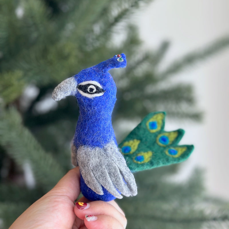 A handcrafted wool felt peacock finger puppet is held by a hand against a dreamy Christmas tree backdrop, showcasing festive charm. This unique needle-felted peacock features a rich blue body with gray accents and a fan of green wool felt tail adorned with blue 'eye' spots. Ethically made, this felt toy for kids brings elegant play.