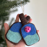A needle felted pickleball ornament is held in a female hand against a festive Christmas tree. The ornament is shaped like a pickleball paddle, featuring a blue upper and teal lower section with a brown edge and handle. A pink felt ball is detailed in the paddle's center. A jute string is attached for hanging.