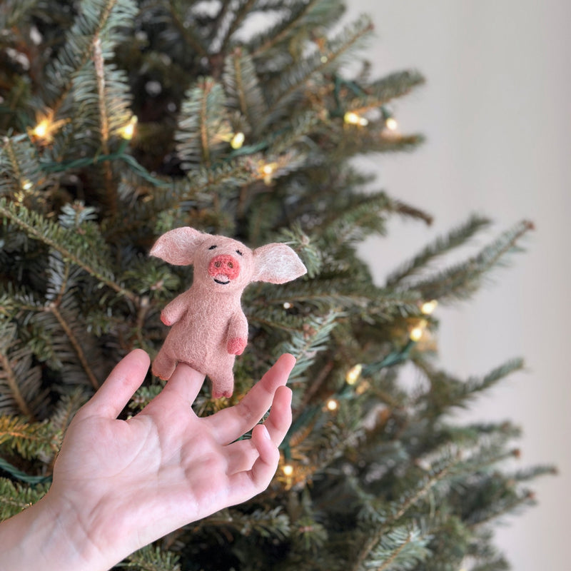 A light pink needle felted pig finger puppet is held up by a female hand, set against a backdrop of a Christmas tree with tiny lights. This lifelike pig features a pink snout, little floppy ears, a black stitched smile, detailed pink hooves, and a cute curly tail.