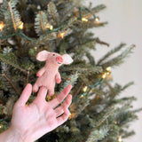 A handmade felt pig finger puppet is held by a female hand, presented against a blurred Christmas tree adorned with delicate lights. This predominantly pink pig features tiny black eyes and a cheerful smile. Its ears and trotters are crafted in a slightly darker pink, adding depth to its cute design.