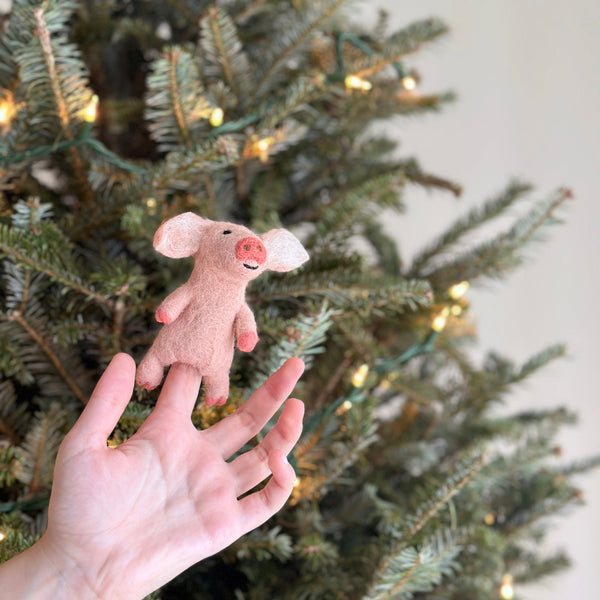 A handmade felt pig finger puppet is held by a female hand, presented against a blurred Christmas tree adorned with delicate lights. This predominantly pink pig features tiny black eyes and a cheerful smile. Its ears and trotters are crafted in a slightly darker pink, adding depth to its cute design.
