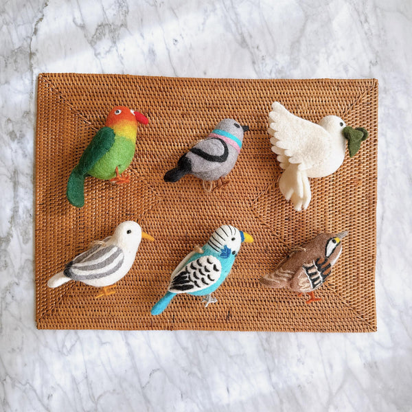 Six felt American bird ornaments rest on a woven wicker mat, including a parrot, a pigeon, a dove with olive twig, a seagull, a blue budgerigar, and a white-throat sparrow. These handmade ornaments feature soft surface, a lifelike shape, and detailed needle-felted textures, complete with small legs. A jute string is attached to each one of the unique Christmas bird ornaments, ready for hanging.