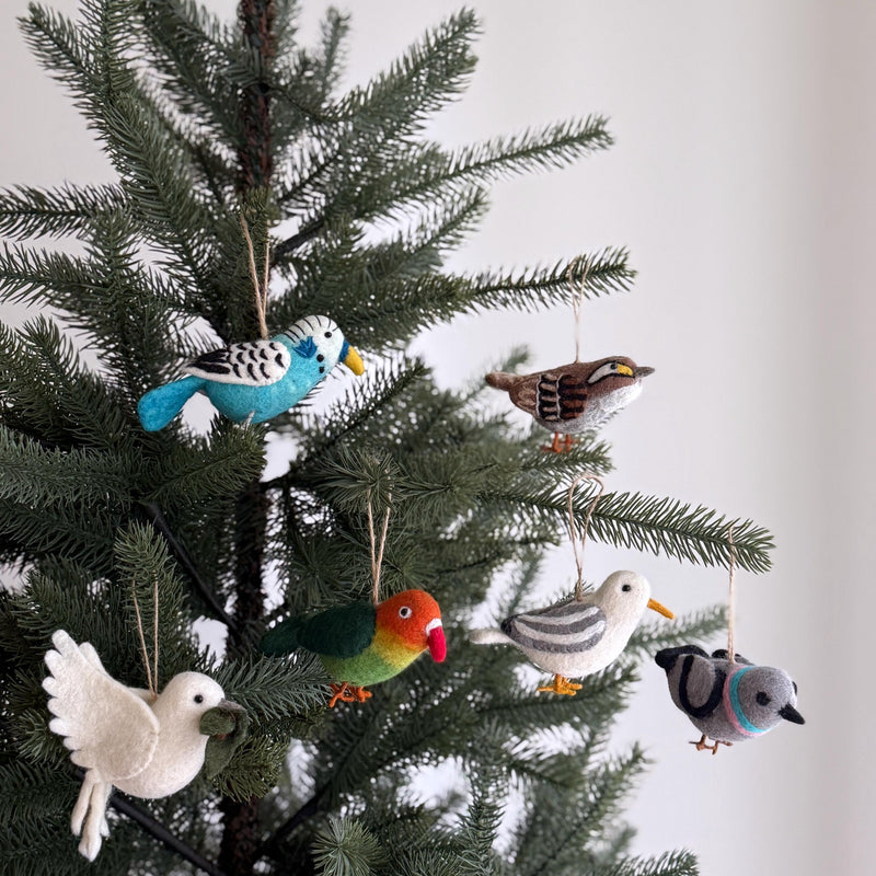 A set of handmade felt bird ornaments hangs on a festive Christmas tree, set against a simple white background. The bird ornament set includes a parrot, a pigeon, a dove with olive twig, a seagull, a blue budgerigar, and a white-throat sparrow. These unique, handcrafted felt ornaments are made from natural wool felt, featuring soft tones and lifelike needle-felted details with visible feather patterns. Each one is suspended by a rustic jute string, which adds a whimsical touch to holiday decor.