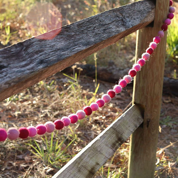 A pink felt ball garland is draped across a weathered wooden fence, creating a warm and inviting outdoor scene with sun-dappled grass and trees in the background. This handmade wool felt garland features alternating felt balls in varying shades of pink, adding a charming touch.