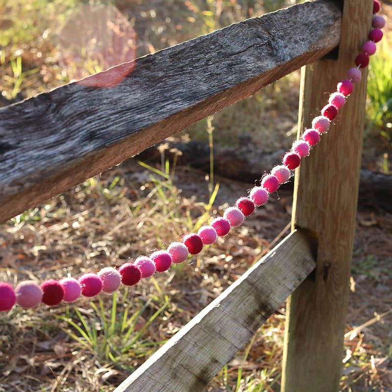 A pink felt ball garland is draped across a weathered wooden fence, creating a warm and inviting outdoor scene with sun-dappled grass and trees in the background. This handmade wool felt garland features alternating felt balls in varying shades of pink, adding a charming touch.