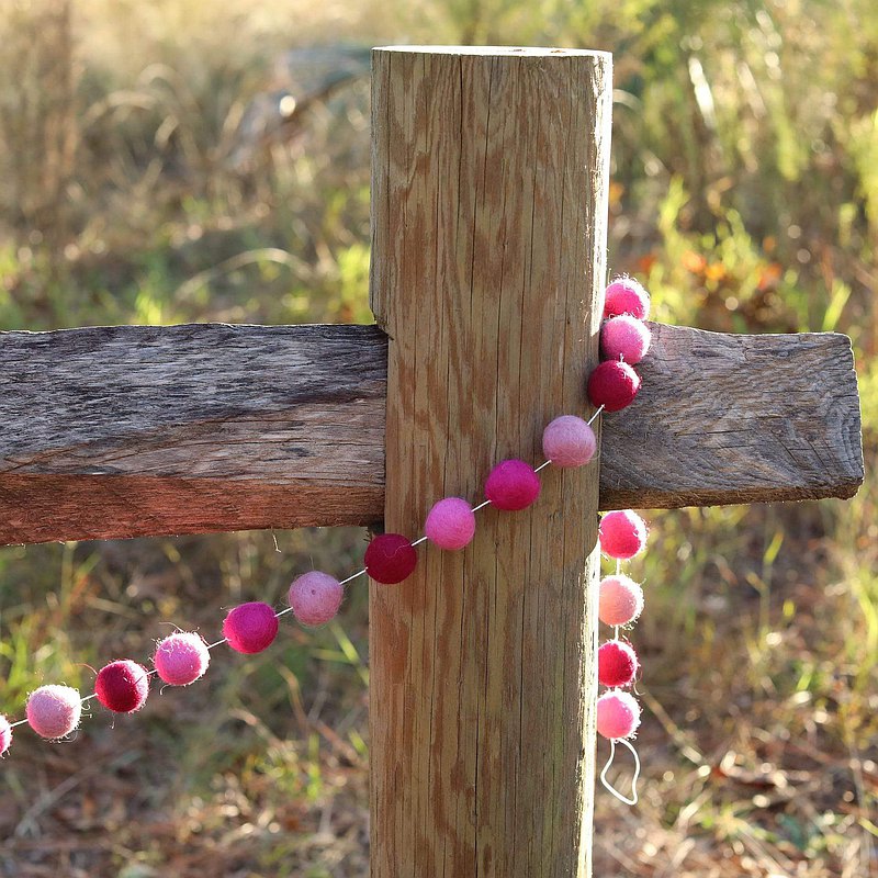 A Pink felt pompom garland is draped whimsically across a weathered wooden fence post, nestled within natural grassy vegetation. The garland showcases round wool felt balls in varying shades of pink, from light blush to deep magenta, evenly spaced to create a symmetrical design, adding to the rustic charm.