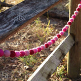 A Pink Felt Ball Garland is draped across a rustic wooden fence, illuminated by natural sunlight. This wool felt garland features round balls in varying shades of pink, from light blush to deep magenta, creating a soft, textured contrast against the weathered wood. Soft grasses and earth tones complete the warm, inviting background.