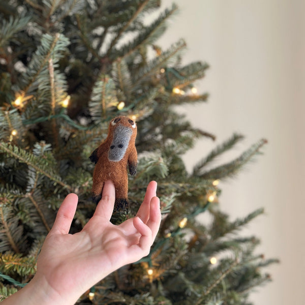 A light-brown, needle-felted platypus finger puppet, crafted from textured wool felt, is gently held by a hand. The charming, handmade felt platypus features a grey bill and small black eyes, positioned against a festive Christmas tree adorned with warm lights. This unique kids finger puppet serves as an ideal storytelling prop or holiday decoration.
