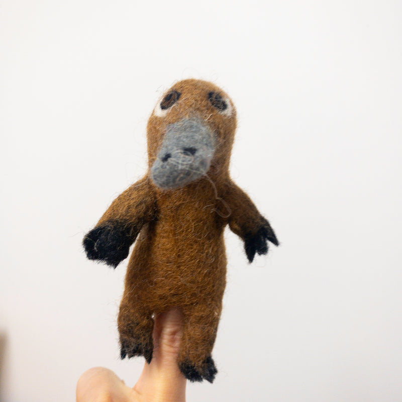 A needle felted wool platypus finger puppet rests on a female finger against a plain white background. The handcrafted felt toy features a brown body, a grey bill, and black feet, highlighting its detailed 3D design.