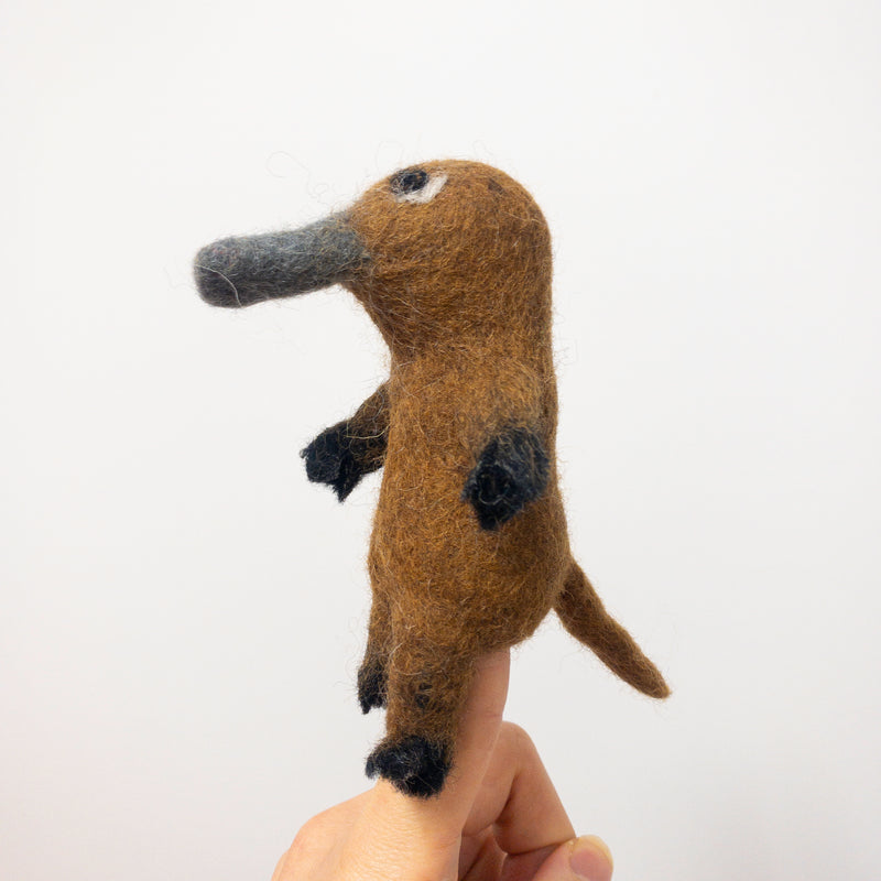 A handcrafted wool felt platypus finger puppet is displayed on a person's finger against a clean white background. This unique needle felted puppet is primarily brown with black accents on its feet and hands. It features a distinctive gray bill and a tail, showcasing its charming 3D-designed shape, ideal as a kids' toy.