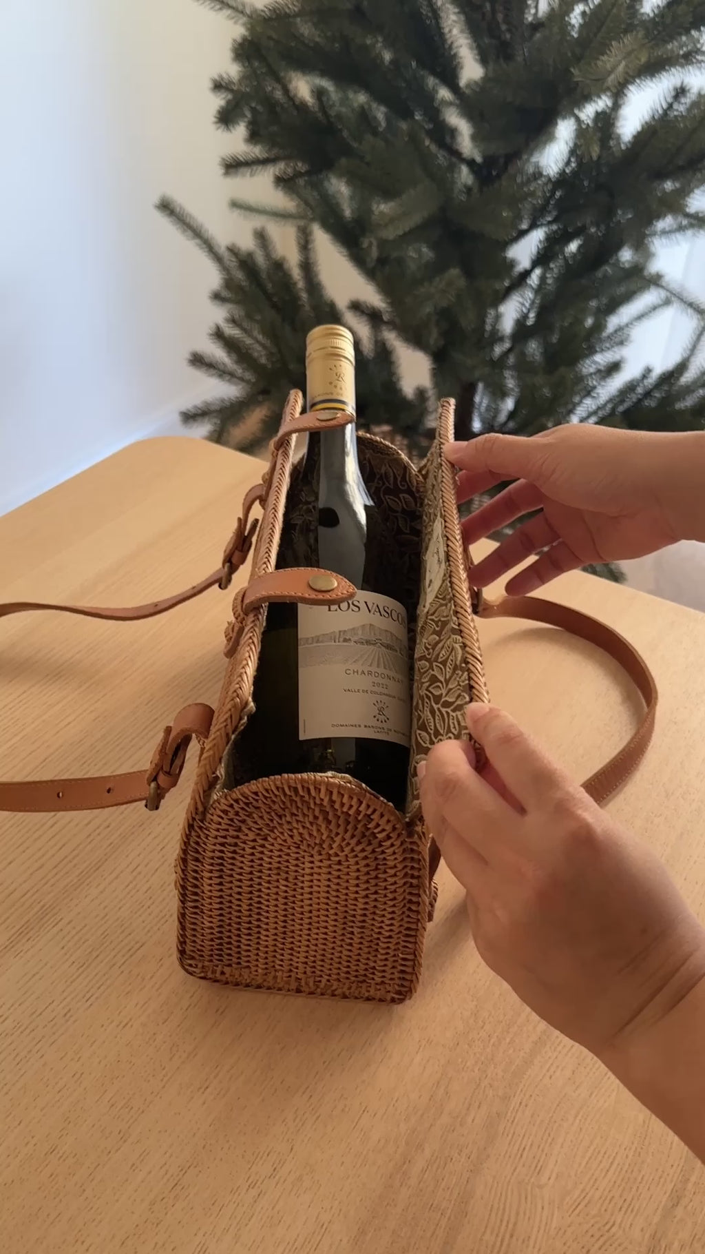 A beautifully crafted Bali rattan handbag, resting on a wooden surface, features a woven rattan material with a unique brown pattern. The bag has an adjustable leather handle and contains a dark-colored wine bottle, suggesting red or black wine. The festive atmosphere, complete with a Christmas tree in the background, implies the handbag is being used as a gift, adding luxury and sophistication to the scene.