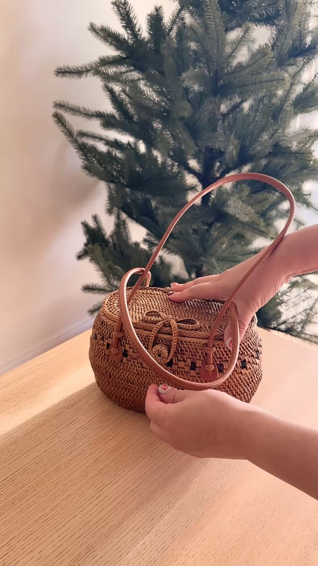 A woven basket, likely a Bali Rattan Bag, is held by a person's hand, resting on a wooden surface with a Christmas tree background. The basket features a copper-colored handle and a woven texture.