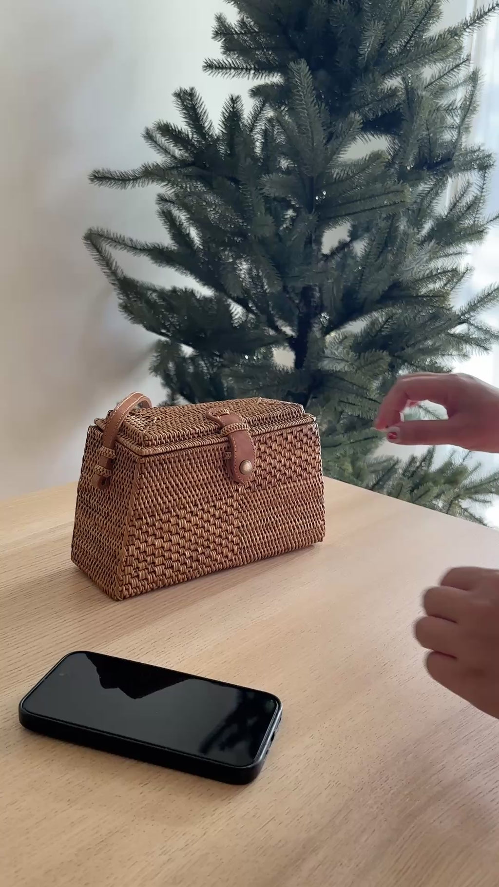 A Bali Rattan Crossbody Bag - Maya rests on a wooden surface, held by a hand at the bottom right corner. The bag features a wicker material with a leather snap clasp, adjustable strap, and unique Batik cotton lining. The festive Christmas tree background conveys comfort and holiday cheer.