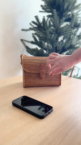 A woven rattan crossbody bag with a leather snap clasp rests on a wooden surface, positioned in front of a Christmas tree. The bag's woven texture and leather details are prominently displayed, showcasing its stylish and durable design.