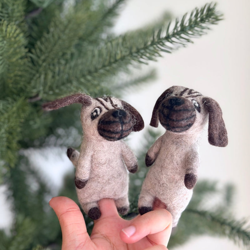 A light brown wool felt pug finger puppet is playfully held by a female hand, set against a blurred Christmas tree background. This charming handmade felt finger puppet features dark brown details on its face, eyes, ears, and mouth, along with small floppy ears and a round body, evoking a cheerful and fun mood.
